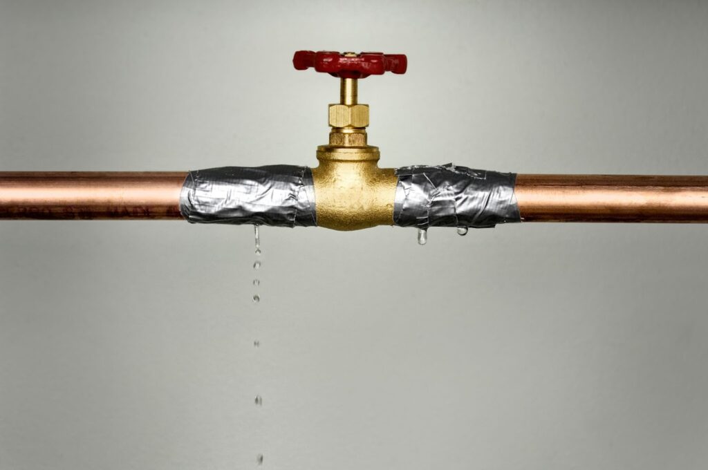 A makeshift repair on a leaking copper pipe with duct tape, failing to stop the drip completely under a closed faucet.