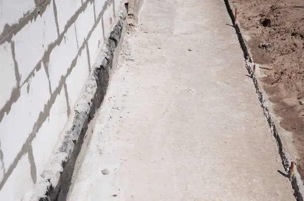 White brick wall with black sealant along the base next to a concrete path and dirt.