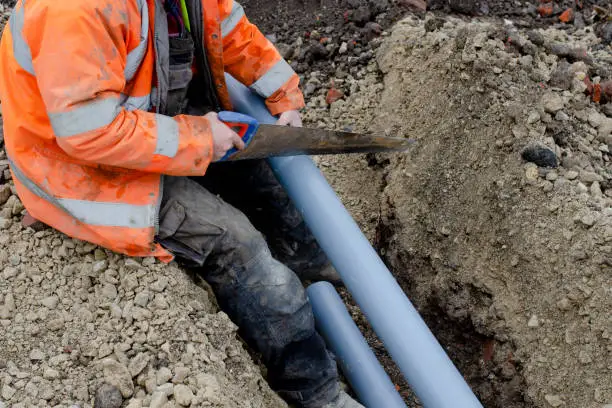 Worker in orange safety vest cutting blue pipes in a trench.