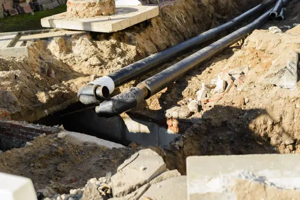 Black pipes with fittings in an excavated trench near a concrete structure.