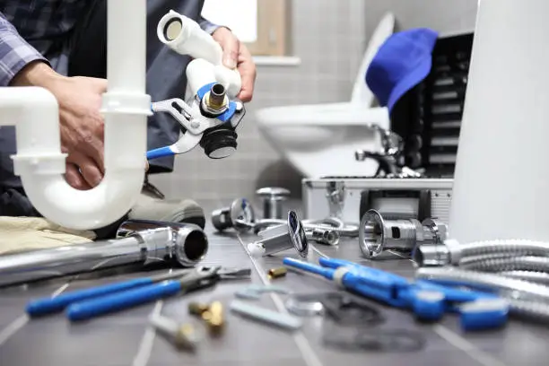 A worker uses a blue-handled wrench on a white pipe, with various plumbing tools and parts scattered on the floor near a toilet.