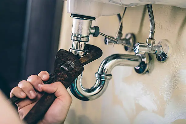 A hand using a rusty wrench to work on a sink's chrome P-trap pipe.